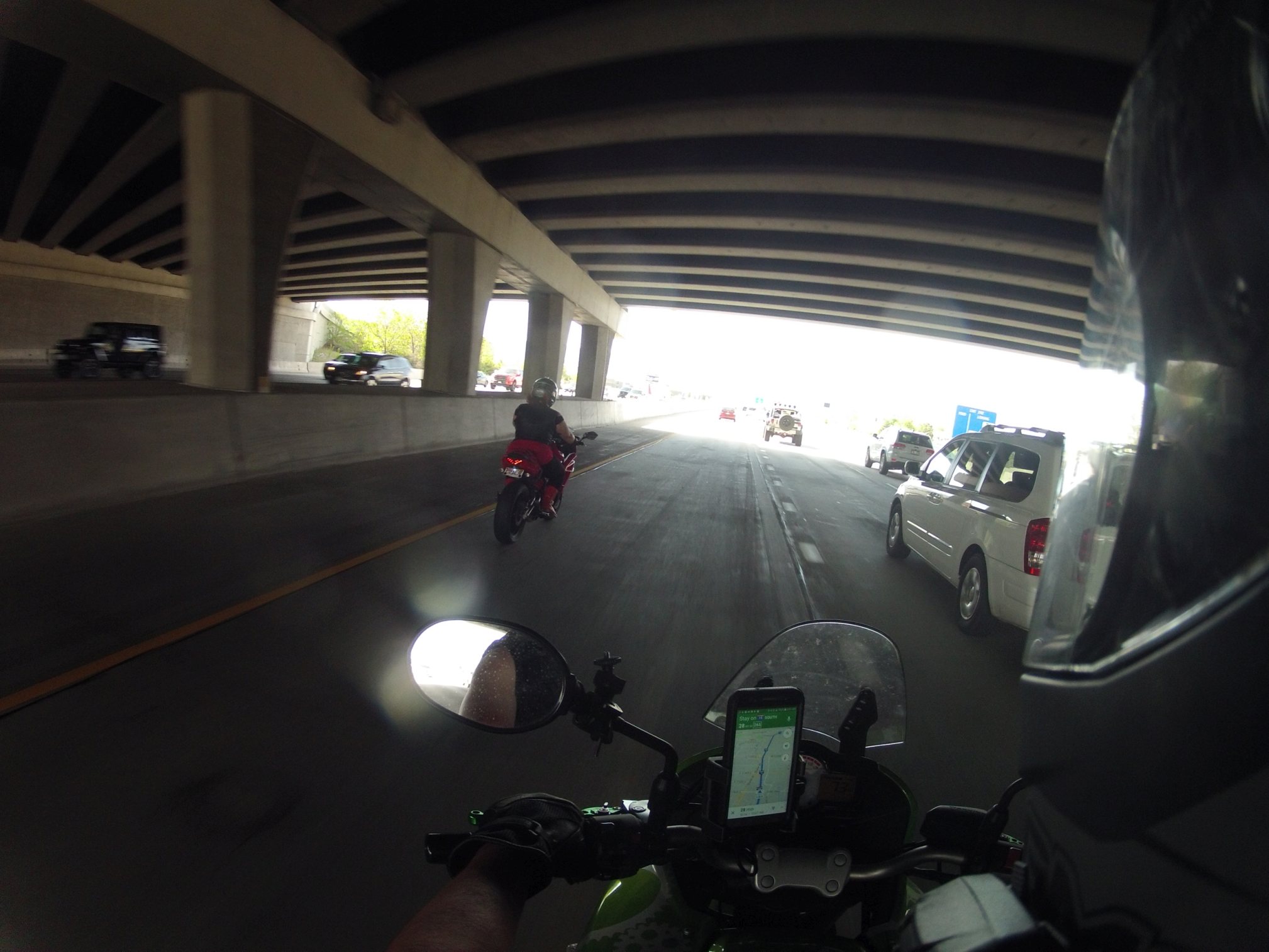 Pictured is a rider astride a motorcycle, helmet visible in the right foreground. Another rider appears two car lengths ahead on the left. They are under a shaded freeway overpass. In the cockpit of the bike a cell phone is visible, displaying GPS navigation with a blue highlighted route.