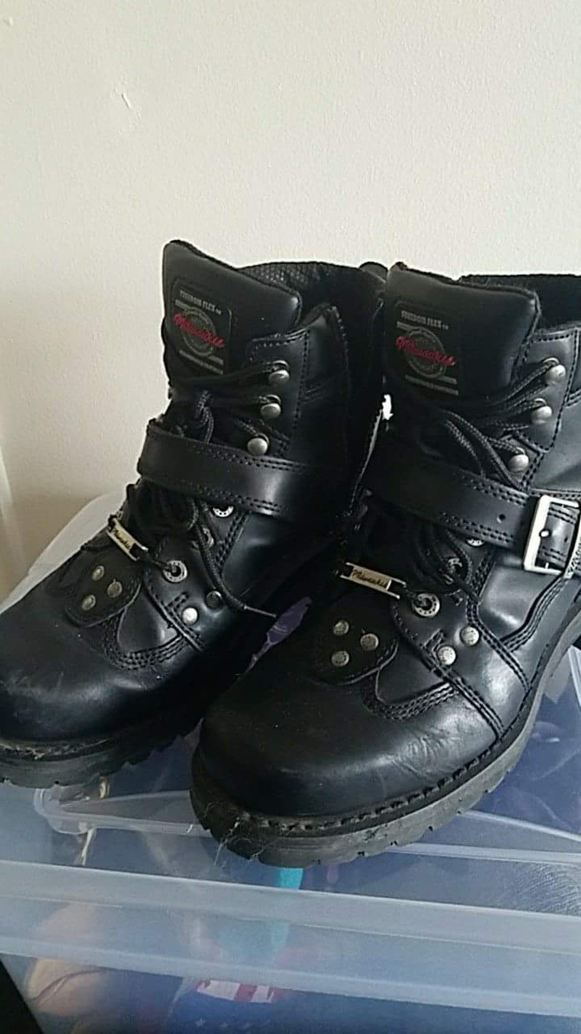 Black shiny leather Milwaukee brand boots with speed lace hooks on the uppers, pictured sitting atop a clear storage bin.