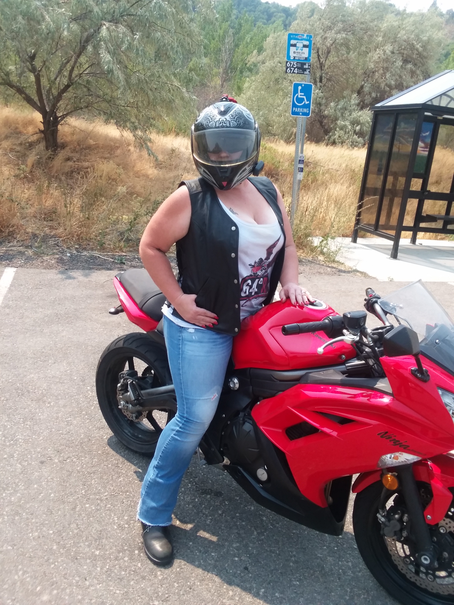 A woman sits astride a red Kawasaki Ninja 650. She is wearing the Rowdy Women's Vest. She wears a white tank top, which displays the image of an aggressive dragon styled after the fairings of a super-sport motorcycle. Behind the woman is a grassy field with scattered trees and a public bus stop.
