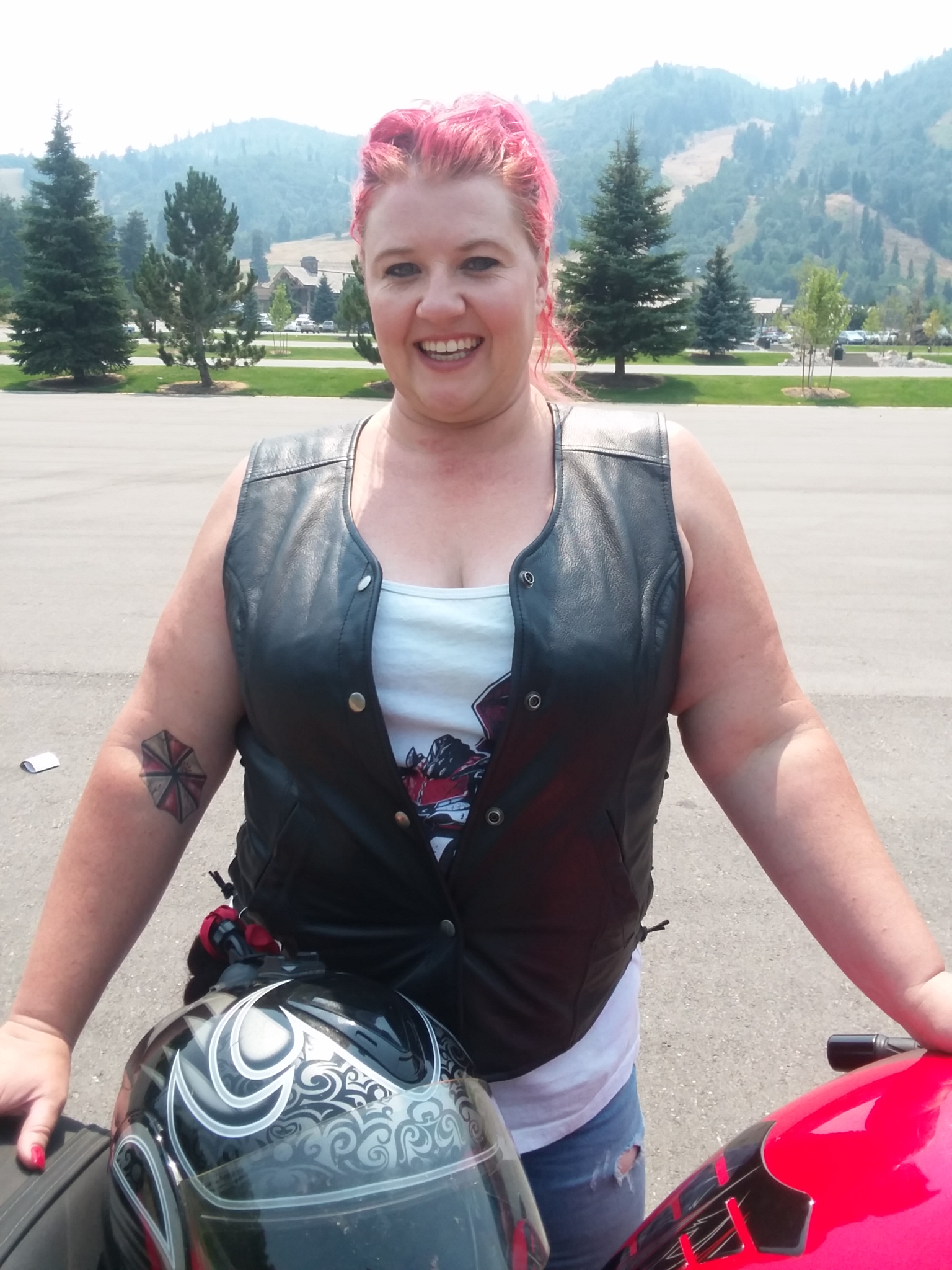 A woman with striking red hair stands next to a Kawasaki Ninja 650. She wears a black leather vest with snaps, undone to her naval, and a white tank top beneath it. An Umbrella Corp logo tattoo is visible in the crook of her right arm. She smiles with good humor. In the background is an expansive parking lot with evergreen trees, and further away, groomed ski slopes rise to the horizon.