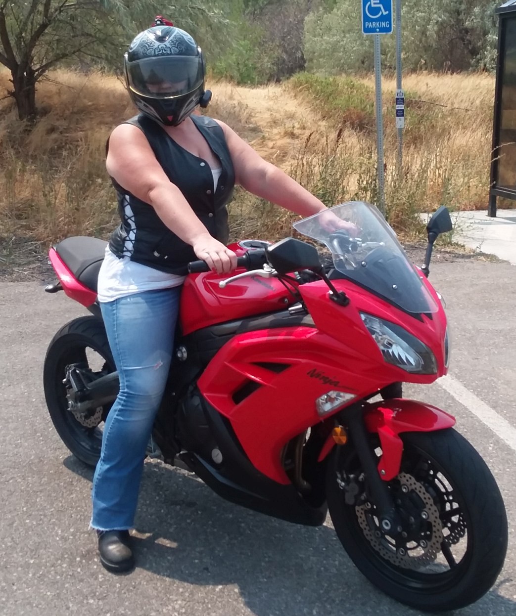 A helmeted woman sits astride a red Kawasaki Ninja 650. Her arms are extended to hold the controls of the bike, and her feet are planted firmly on the ground. She wears black boots, blue jeans, a white tank top and a black leather vest. The vest is partially unsnapped at the neckline to show the white top beneath. In the background, dry grass and scattered trees and a public bus stop.