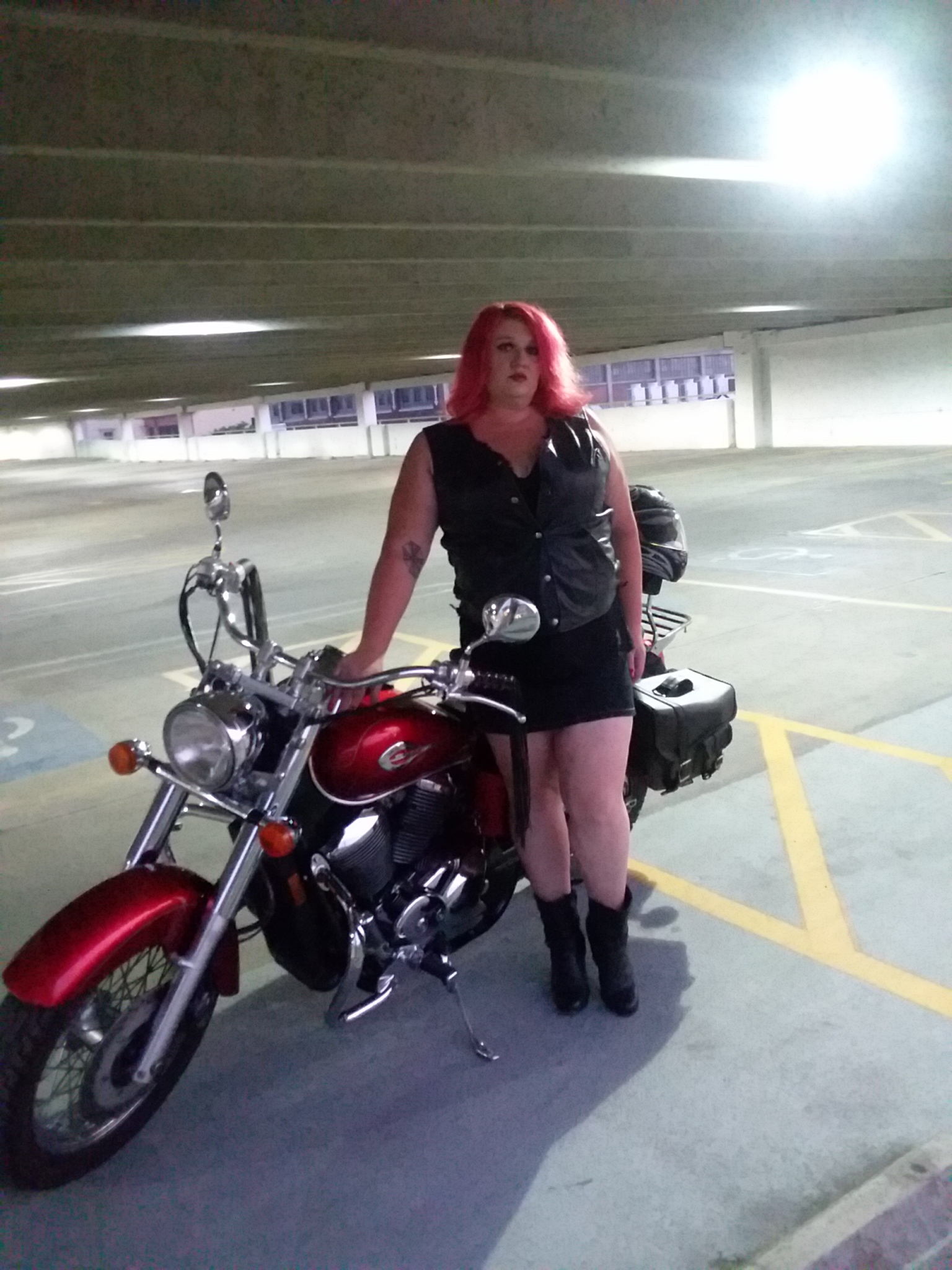 A woman with striking red hair stands beside a polished Honda Shadow ACE in a concrete parking garage. She wear a leather vest snapped closed, a skirt and high heeled boots. Above and to her right, a bright light bathes the scene with intense white illumination.