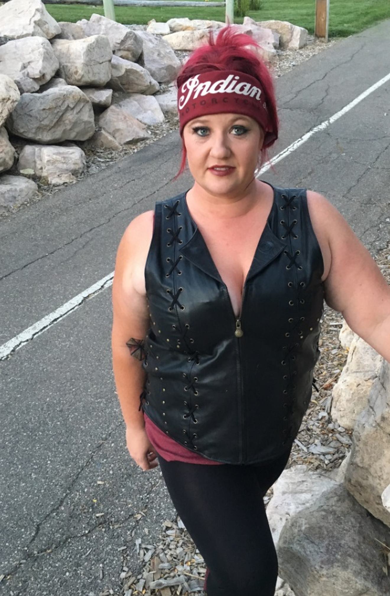 Indian Women's Review, a woman stands next to a rock wall, she is wearing a a black leather motorcycle vest and a Bandana around her forehead showing the Indian Logo. The color bandanna matches the women's vibrant dark red hair.
