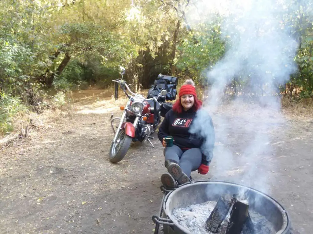 Indian Women's Review, A smiling woman holds a steaming cup of coffee, she sits with her feet up next to a smoking campfire in the woods. She is wearing a black hoodie with the 64 degree racing logo and a red knit hat and fingerless gloves. Her shiny red motorcycle is sitting behind her stacked with an assortment of black leather motorcycle luggage.