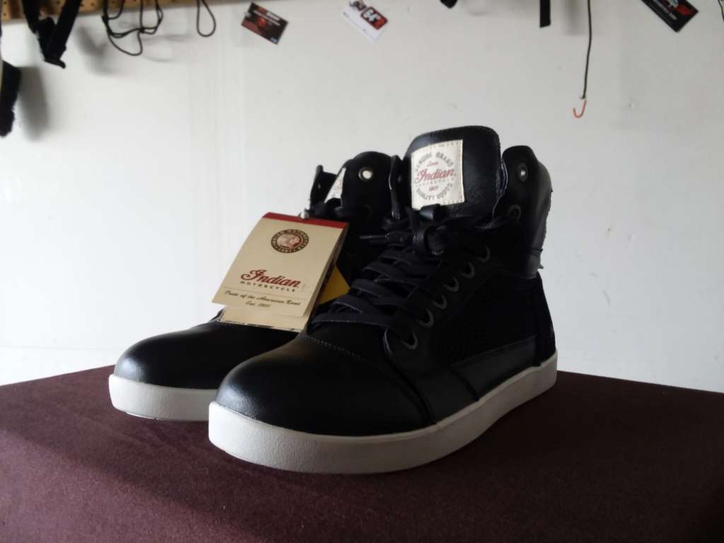 The Mesh Hi-Top Sneakers from Indian sit casually on a brown surface, a garage wall with manufacturer decals in the background. There is stark contrast between the white soles and black uppers.