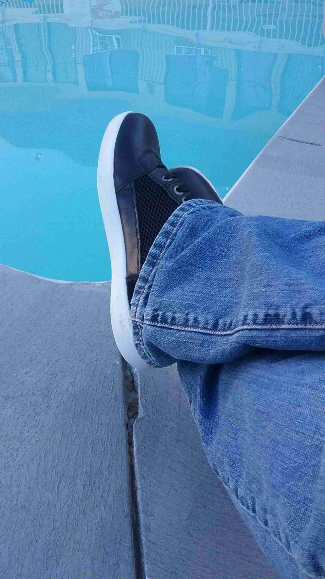 A man's feet, crossed at the ankles, is shown in the foreground. He's wearing the Mesh Hi-Top Sneakers from Indian Motorcycle, and they appear comfortable and relaxed. In front of him is the corner of a motel pool, the water inviting and blue.