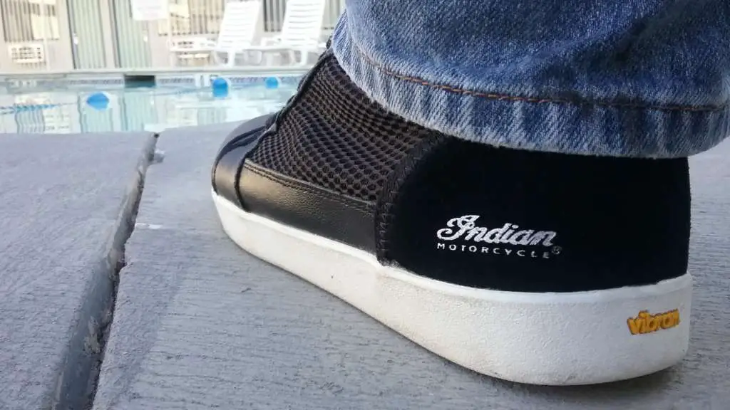An extreme closeup of a Mesh Hi-Top Sneaker from Indian Motorcycle, showing the logo on the heel and the bright white sole stretching away. The mesh panel is clearly visible as well. In the background, a motel pool with desk chairs is vaguely visible.