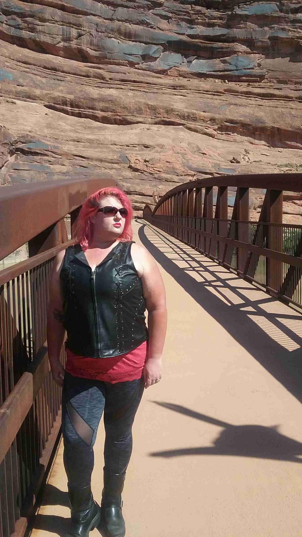 Indian Women's Review, A woman with striking red hair stands on a long river bridge in front of a beautiful red rock mountain wall, she is wearing a black leather motorcycle vest, motorcycle boots and sunglasses.