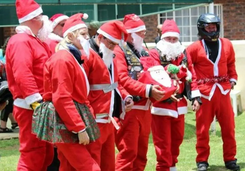 7 men dressed in Santa suits and beards. Several are holding gifts. One santa has his coat duct taped shut and one Santa has a tinsel belt and is wearing a motorcycle helmet! 