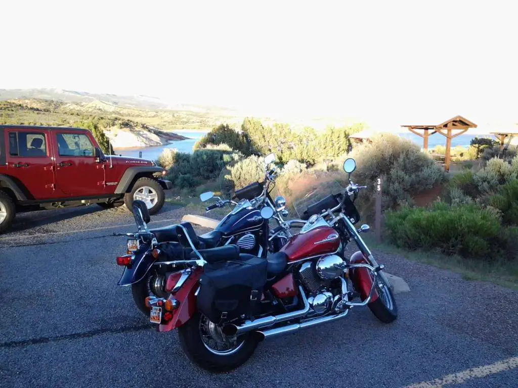 Red Honda Shadow ACE and blue Kawasaki Vulcan parked next to a campsite and large reservoir.