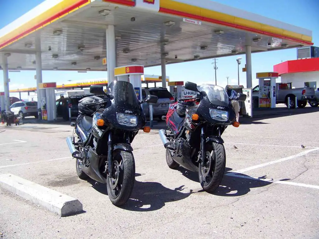 Two identical Kawasaki Ninja 500EX's parked on the apron at a large fuel plaza.