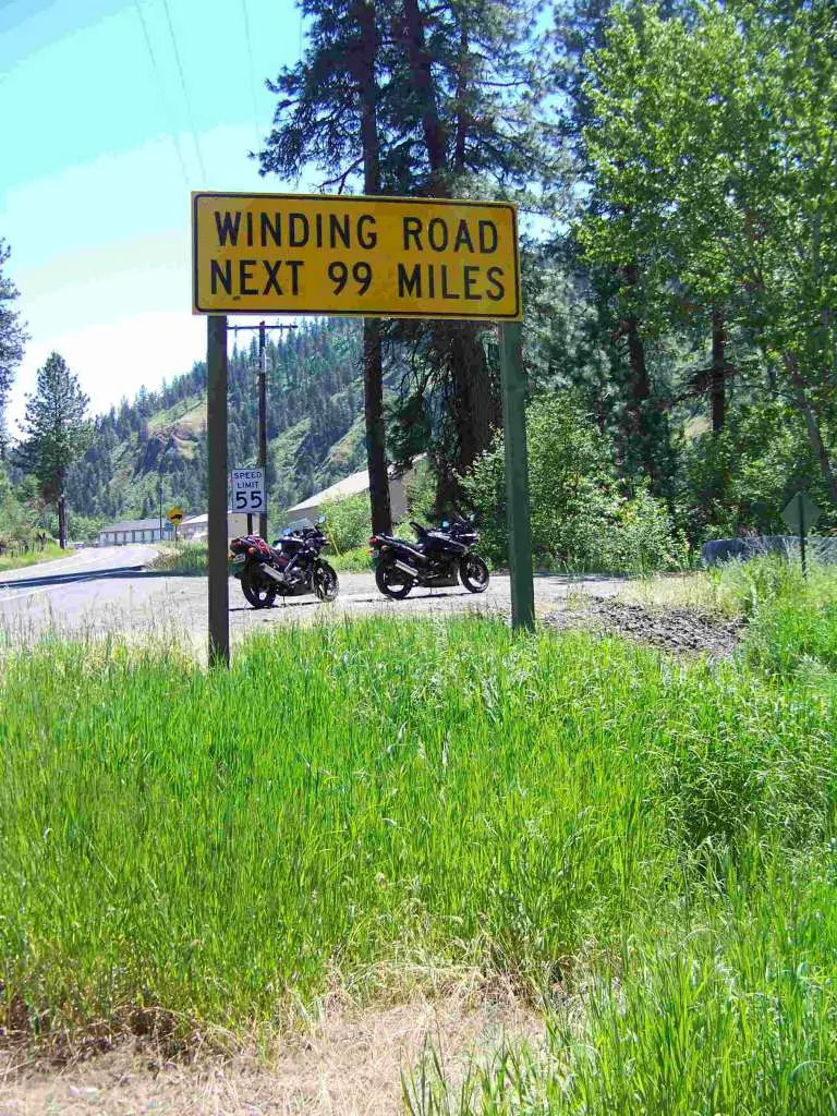 Identical Kawasaki Ninja 500EX's are pictured on a gravel shoulder of a narrow mountain road. Prominent in the foreground, a signpost states "WINDING ROAD NEXT 99 MILES".