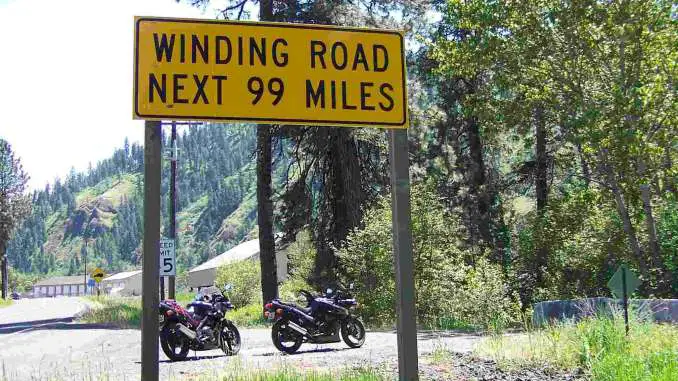 Two black sport motorcycles are parked in a forest near a yellow road sign that reads 'Winding Road Next 99 Miles'