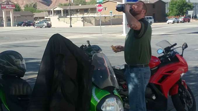 A man takes a large gulp of soda from a bottle with one hand, a gas station taquito in the other. He is standing between two Kawasaki Sport motorcycles in a 7-11 gas station parking lot. The barely visible background scenery includes beautiful red cliffs.