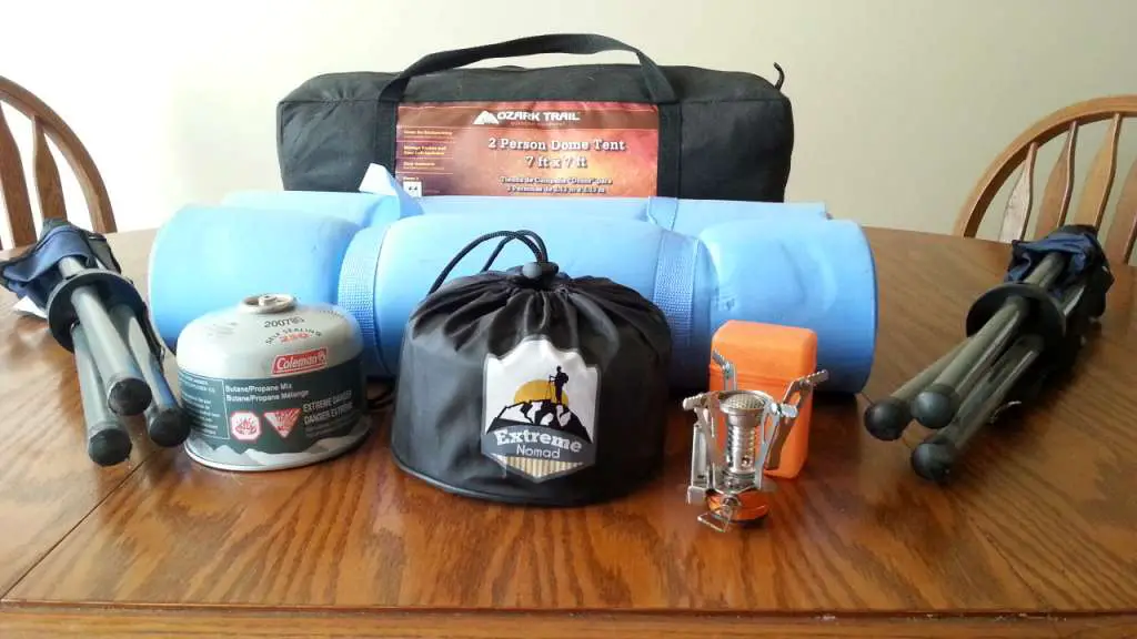 Various Camping Gear Including a compact dome tent, 2 bed rolls, 2 fold up tripod camp chairs, a butane fuel bottle, a travel size burner and a compact mess kit set atop a table.