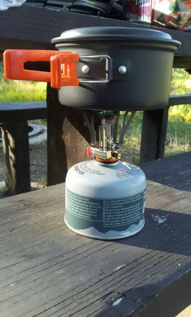 A Butane burner is placed atop a camp table with a mess kit pot heating on the burner.