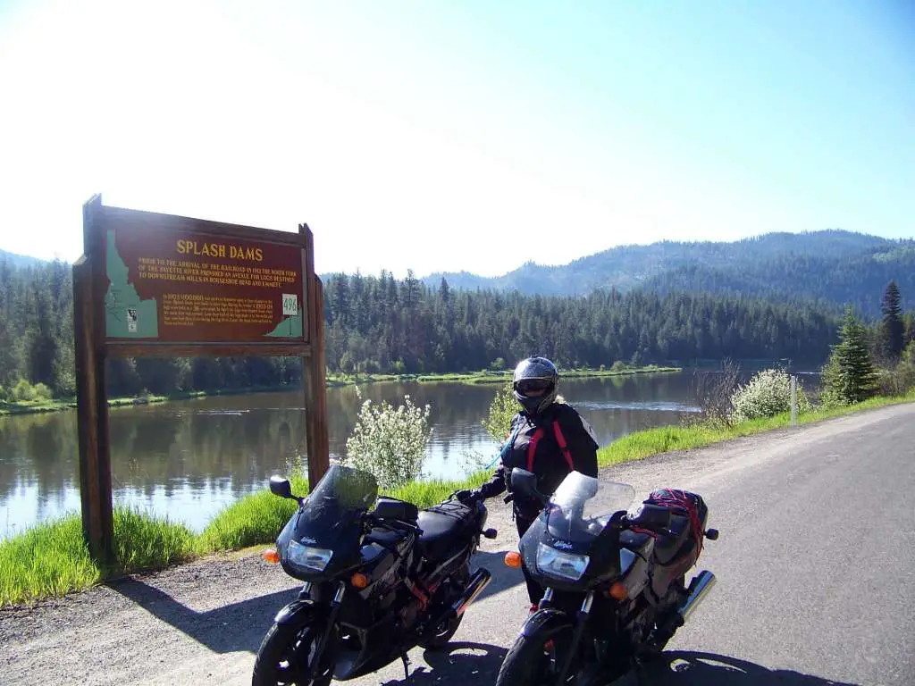 A girl in full motorcycle gear stands between two sport motorcycles next to a beautiful river with mountains and forest on the other sign. A sign explains 'Splash Dams'.