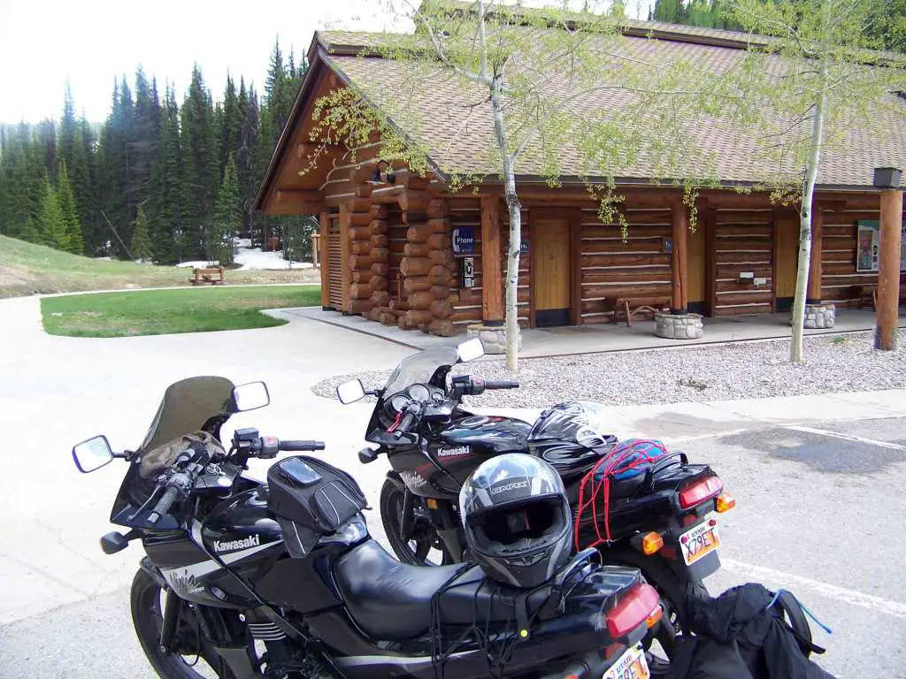 Two black sport motorcycles are parked near a log cabin club house in a forest with a little bit of snow under the trees. The parking lot is dry and the sun is out.