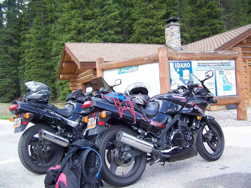 Two black sport motorcycles are parked together in a parking lot in a forest. The background shows an Idaho map and a Log Cabin Visitor's Center. 