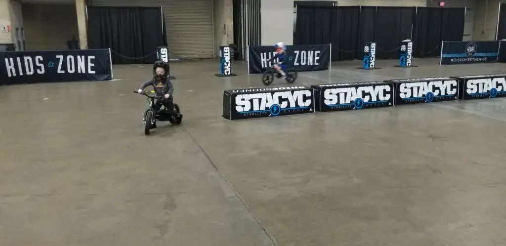 Young children in full motorcycle gear navigate a closed, padded oval track on STACYC Stability bikes.